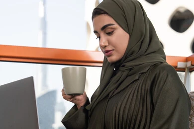 Busy working, enjoying a cup of hot drink, young veiled Emirati Gulf Arab woman wearing green abaya and hijab, working remotely using internet via laptop in an outdoor high-rise cafe