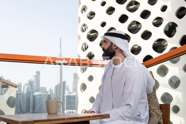 Enjoying spending free time outdoors, entertainment and fun during the holiday, a side view of an Emirati Gulf Arab man wearing a kandura and a white ghutrah sitting in a café contemplating the natural scenery.
