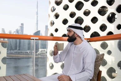 تناول الأعشاب الشتوية الدافئة, تعديل المزاج بشرب الكافيئين,  صورة من الجانب لرجل عربي خليجي اماراتي يرتدي الكندورة و الغتره البيضاء يجلس فى المقهي و يمسك كوب القهوه , الاستمتاع بقضاء وقت الفراغ فى الخارج