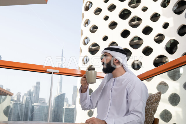 الاستمتاع بقضاء وقت الفراغ فى الخارج, تعديل المزاج بشرب الكافيئين، الاستمتاع بتناول المشروبات الساخنة, صورة من الجانب لرجل عربي خليجي اماراتي يرتدي الكندورة و الغتره البيضاء يجلس فى المقهي و يمسك كوب القهوه