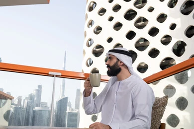 Enjoying spending free time outdoors, improving mood by drinking caffeine, enjoying hot beverages, a side view of an Arab Gulf Emirati man wearing a kandura and a white ghutrah sitting in a café holding a cup of coffee.