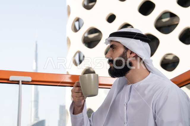 الاستمتاع بتناول المشروبات الساخنة, تعديل المزاج بشرب الكافيئين, صورة مقربة من الجانب لرجل عربي خليجي اماراتي يرتدي الكندورة و الغتره البيضاء يجلس فى المقهي و يمسك كوب القهوه , الاستمتاع بقضاء وقت الفراغ فى الخارج