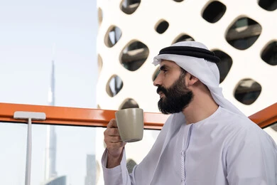 Enjoying hot beverages, mood enhancement by drinking caffeine, a close-up side view of an Emirati Gulf Arab man wearing a kandura and a white ghutrah sitting in a café holding a cup of coffee, enjoying spending leisure time outdoors.