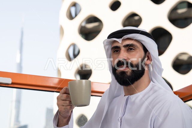 تعديل المزاج بشرب الكافيئين، الاستمتاع بتناول المشروبات الساخنة, صورة مقربة لرجل عربي خليجي اماراتي يرتدي الكندورة و الغتره البيضاء يجلس فى المقهي و يمسك كوب القهوه , الاستمتاع بقضاء وقت الفراغ فى الخارج , ايماءات البشاشة و السرور