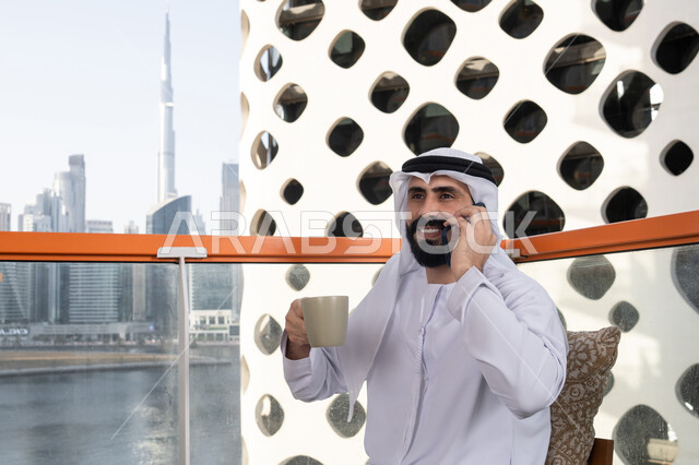 Communicating with family and friends, making a phone call, spending enjoyable time outdoors, a young Emirati Gulf Arab man wearing a kandura and a white ghutrah sitting in a café, the concept of technology and using modern techniques, remote communication, conducting a live broadcast, enjoying hot beverages.