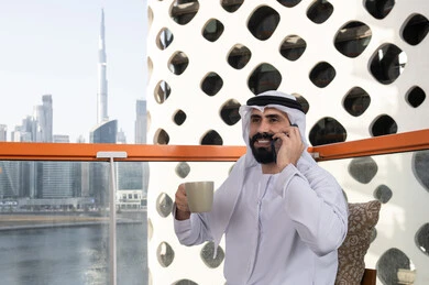 Communicating with family and friends, making a phone call, spending enjoyable time outdoors, a young Emirati Gulf Arab man wearing a kandura and a white ghutrah sitting in a café, the concept of technology and using modern techniques, remote communication, conducting a live broadcast, enjoying hot beverages.