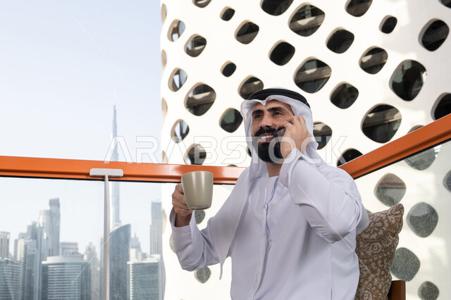Live broadcast, making a phone call, having a good time outdoors, a young Arab Gulf Emirati man wearing a kandura and a white ghutrah sitting in a café, the concept of technology and using modern techniques, remote communication, connecting with family and friends, enjoying hot beverages.