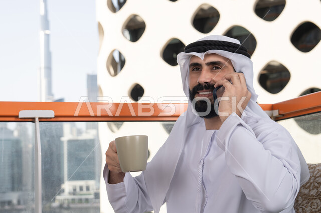 التواصل عن بعد,اجراء مكالمه هاتفيه، قضاء وقت ممتع فى الخارج ،صورة مقربة  لشاب عربي خليجي إماراتي يرتدي الكندورة والغترة البيضاء يجلس في المقهى ، مفهوم التكنولوجيا وإستخدام تقنيات حديثة, اجراء بث مباشر,التواصل مع الاهل والاصدقاء, الاستمتاع بتناول المشروبات الساخنه