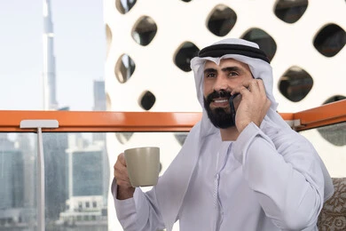 Remote communication, making a phone call, having a good time outdoors, a close-up image of a young Emirati Arab man wearing a kandura and a white ghutrah sitting in a café, the concept of technology and using modern techniques, conducting a live broadcast, communicating with family and friends, enjoying hot beverages.