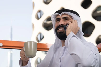 الاستمتاع بالمشروبات الساخنه,اجراء مكالمه هاتفيه، قضاء وقت ممتع فى الخارج ، صورة مقربة لشاب عربي خليجي إماراتي يرتدي الكندورة والغترة البيضاء يجلس في المقهى ، مفهوم التكنولوجيا وإستخدام تقنيات حديثة, التواصل عن بعد, اجراء بث مباشر,التواصل مع الاهل والاصدقاء