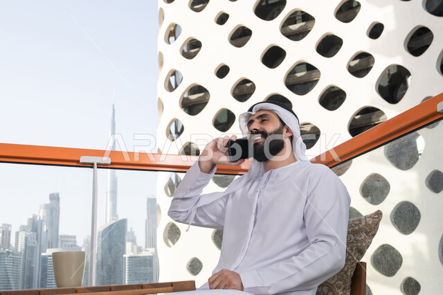 مفهوم التكنولوجيا وإستخدام تقنيات حديثة. اجراء مكالمه هاتفيه، قضاء وقت ممتع فى الخارج ، شاب عربي خليجي إماراتي يرتدي الكندورة والغترة البيضاء يجلس في المقهى , التواصل عن بعد, اجراء بث مباشر,التواصل مع الاهل والاصدقاء, الاستمتاع بالمشروبات الساخنه