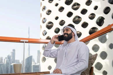 The concept of technology and the use of modern techniques. Making a phone call, spending enjoyable time outdoors, a young Arab Gulf Emirati man wearing a kandura and a white ghutrah sitting in a café, remote communication, conducting a live broadcast, communicating with family and friends, enjoying hot beverages.