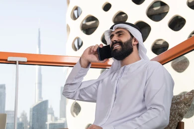 A close-up image of a young Emirati Arab man wearing a kandura and a white ghutrah sitting in a café, making a phone call, enjoying time outdoors, the concept of technology and using modern techniques, remote communication, conducting a live broadcast, connecting with family and friends.