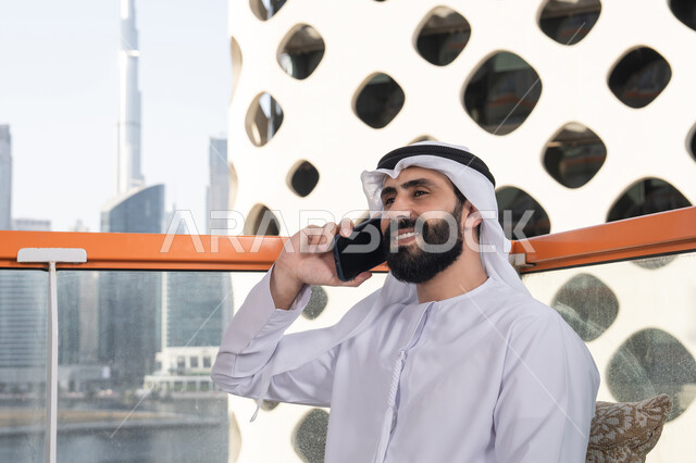 Spending enjoyable time outdoors, making a phone call, a close-up image of a young Arab Gulf Emirati man wearing a kandura and a white ghutrah sitting in a café, the concept of technology and the use of modern techniques, remote communication, conducting a live broadcast, communicating with family and friends.