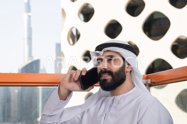 اجراء مكالمه هاتفيه، قضاء وقت ممتع فى الخارج ،صورة مقربة لشاب عربي خليجي إماراتي يرتدي الكندورة والغترة البيضاء يجلس في المقهى ، مفهوم التكنولوجيا وإستخدام تقنيات حديثة, التواصل عن بعد, اجراء بث مباشر,التواصل مع الاهل والاصدقاء