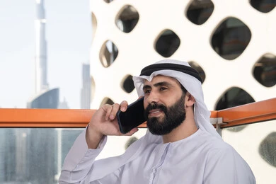 Making a phone call, having a good time outdoors, a close-up image of a young Emirati Arab man wearing a kandura and a white ghutrah sitting in a café, the concept of technology and the use of modern techniques, remote communication, conducting a live broadcast, communicating with family and friends.