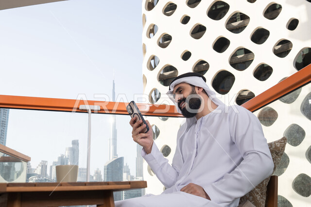 الاستمتاع بقضاء العطله في الخارج, ايماءات السعادة و السرور، تصفح التطبيقات وبرامج التواصل الاجتماعي، التواصل وتبادل الأحاديث والأخبار مع الأهل والأصدقاء، رجل عربي خليجي اماراتي يرتدي الكندورة والغترة البيضاء يجلس فى المقهي و يمسك بيده الهاتف المحمول، استخدام جهاز تقني حديث ومتطور