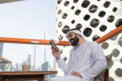 Enjoying spending the holiday abroad, gestures of happiness and joy, browsing applications and social media programs, communicating and exchanging conversations and news with family and friends, an Arab Gulf Emirati man wearing the kandura and white ghutrah sitting in a café holding a mobile phone in his hand, using a modern and advanced technological device.
