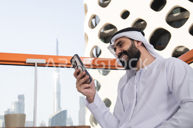 قضاء وقت ممتع عبر الجوال مع الأصحاب,ايماءات السعادة و السرور، تصفح التطبيقات وبرامج التواصل الاجتماعي، التواصل وتبادل الأحاديث والأخبار مع الأهل والأصدقاء، صورة مقربة لرجل عربي خليجي اماراتي يرتدي الكندورة والغترة البيضاء يجلس فى المقهي و يمسك بيده الهاتف المحمول، استخدام جهاز تقني حديث ومتطور