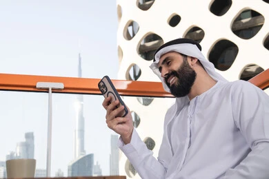 قضاء وقت ممتع عبر الجوال مع الأصحاب,ايماءات السعادة و السرور، تصفح التطبيقات وبرامج التواصل الاجتماعي، التواصل وتبادل الأحاديث والأخبار مع الأهل والأصدقاء، صورة مقربة لرجل عربي خليجي اماراتي يرتدي الكندورة والغترة البيضاء يجلس فى المقهي و يمسك بيده الهاتف المحمول، استخدام جهاز تقني حديث ومتطور