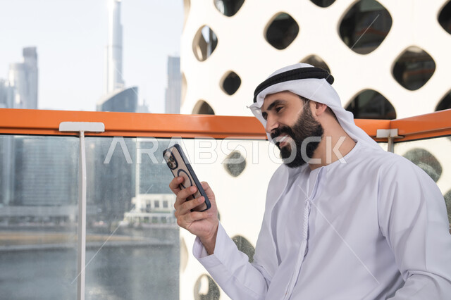 تعابير السرور والاندماج, العمل عن بعد، تصفح التطبيقات وبرامج التواصل الاجتماعي، التواصل وتبادل الأحاديث والأخبار مع الأهل والأصدقاء، صورة مقربة لرجل عربي خليجي اماراتي يرتدي الكندورة والغترة البيضاء يجلس فى المقهي و يمسك بيده الهاتف المحمول، استخدام جهاز تقني حديث ومتطور