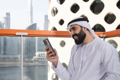 تعابير السرور والاندماج, العمل عن بعد، تصفح التطبيقات وبرامج التواصل الاجتماعي، التواصل وتبادل الأحاديث والأخبار مع الأهل والأصدقاء، صورة مقربة لرجل عربي خليجي اماراتي يرتدي الكندورة والغترة البيضاء يجلس فى المقهي و يمسك بيده الهاتف المحمول، استخدام جهاز تقني حديث ومتطور