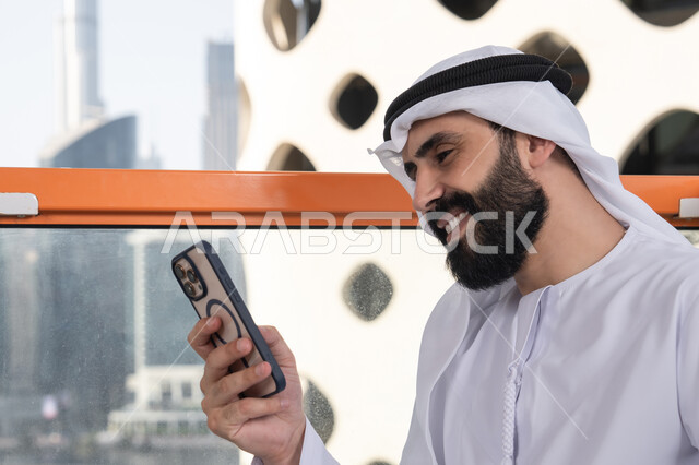 Browsing the internet and using modern technologies, gestures of happiness and joy, remote work, communication and exchanging conversations and news with family and friends, a close-up image of an Emirati Gulf Arab man wearing a kandura and a white ghutrah sitting in a café holding a mobile phone, using a modern and advanced technological device.
