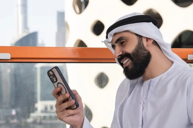 تصفح الانترنت واستخدام تقنيات حديثة, ايماءات السعادة و السرور, العمل عن بعد، التواصل وتبادل الأحاديث والأخبار مع الأهل والأصدقاء،  صورة مقربة لرجل عربي خليجي اماراتي يرتدي الكندورة والغترة البيضاء يجلس فى المقهي و يمسك بيده الهاتف المحمول، استخدام جهاز تقني حديث ومتطور
