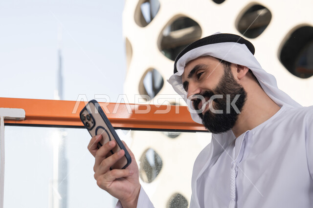 ايماءات السعادة و السرور, العمل عن بعد، تصفح التطبيقات وبرامج التواصل الاجتماعي، التواصل وتبادل الأحاديث والأخبار مع الأهل والأصدقاء، صورة مقربة لرجل عربي خليجي اماراتي يرتدي الكندورة والغترة البيضاء يجلس فى المقهي و يمسك بيده الهاتف المحمول، استخدام جهاز تقني حديث ومتطور