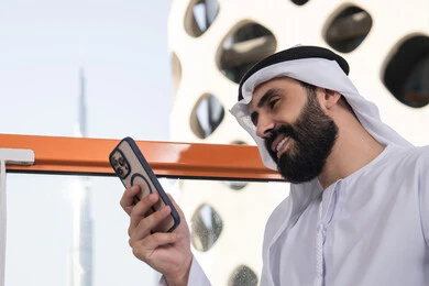 Gestures of happiness and joy, remote work, browsing applications and social media programs, communicating and exchanging conversations and news with family and friends, a close-up image of an Emirati Gulf Arab man wearing a kandura and a white ghutrah sitting in a café holding a mobile phone, using a modern and advanced technological device.