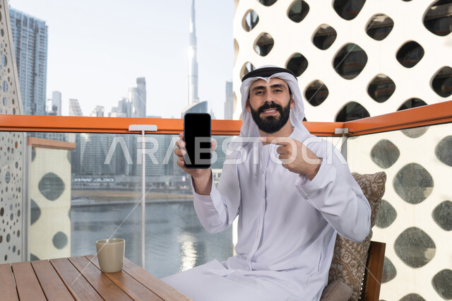 الاستمتاع بتناول المشروبات, عرض شاشة سوداء فارغة على الهاتف المحمول، استخدام الوسائل التقنية الحديثة، النظر إلى الكاميرا بايماءات الفرح والسرور، رجل عربي خليجي اماراتي مبتسم يرتدي الكندورة و الغتره يجلس في المقهي يحمل الجوال ويشير اليه، قضاء وقت ممتع فى الخارج
