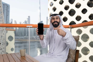 الاستمتاع بتناول المشروبات, عرض شاشة سوداء فارغة على الهاتف المحمول، استخدام الوسائل التقنية الحديثة، النظر إلى الكاميرا بايماءات الفرح والسرور، رجل عربي خليجي اماراتي مبتسم يرتدي الكندورة و الغتره يجلس في المقهي يحمل الجوال ويشير اليه، قضاء وقت ممتع فى الخارج