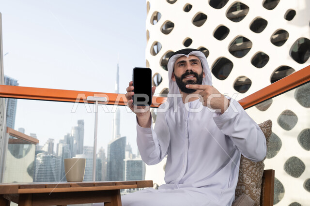 استخدام التكنولوجيا المتطورة للتسويق الرقمي الإلكتروني, عرض شاشة سوداء فارغة على الهاتف المحمول، النظر إلى الكاميرا بايماءات الفرح والسرور، رجل عربي خليجي اماراتي مبتسم يرتدي الكندورة و الغتره يجلس في المقهي يحمل الجوال ويشير اليه، قضاء وقت ممتع فى الخارج