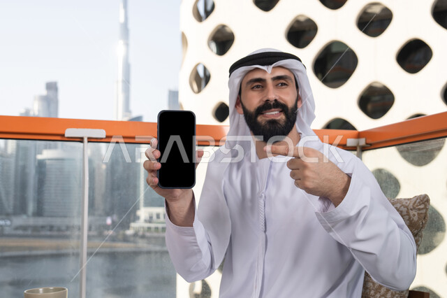 تطبيقات وبرامج الجوال والتسوق الالكتروني, عرض شاشة سوداء فارغة على الهاتف المحمول، استخدام الوسائل التقنية الحديثة، النظر إلى الكاميرا بايماءات الفرح والسرور، رجل عربي خليجي اماراتي مبتسم يرتدي الكندورة و الغتره يجلس في المقهي يحمل الجوال ويشير اليه، قضاء وقت ممتع فى الخارج