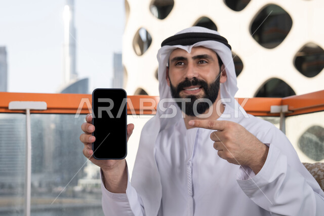قضاء وقت ممتع فى الخارج,عرض شاشة سوداء فارغة على الهاتف المحمول، استخدام الوسائل التقنية الحديثة، النظر إلى الكاميرا بايماءات الفرح والسرور، رجل عربي خليجي اماراتي مبتسم يرتدي الكندورة و الغتره يجلس في المقهي يحمل الجوال ويشير اليه