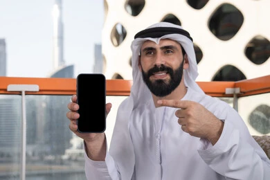 قضاء وقت ممتع فى الخارج,عرض شاشة سوداء فارغة على الهاتف المحمول، استخدام الوسائل التقنية الحديثة، النظر إلى الكاميرا بايماءات الفرح والسرور، رجل عربي خليجي اماراتي مبتسم يرتدي الكندورة و الغتره يجلس في المقهي يحمل الجوال ويشير اليه