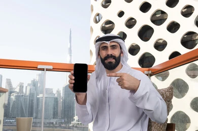رجل عربي خليجي اماراتي مبتسم يرتدي الكندورة و الغتره يجلس في المقهي يحمل الجوال ويشير اليه, عرض شاشة سوداء فارغة على الهاتف المحمول، استخدام الوسائل التقنية الحديثة، النظر إلى الكاميرا بايماءات الفرح والسرور، قضاء وقت ممتع فى الخارج