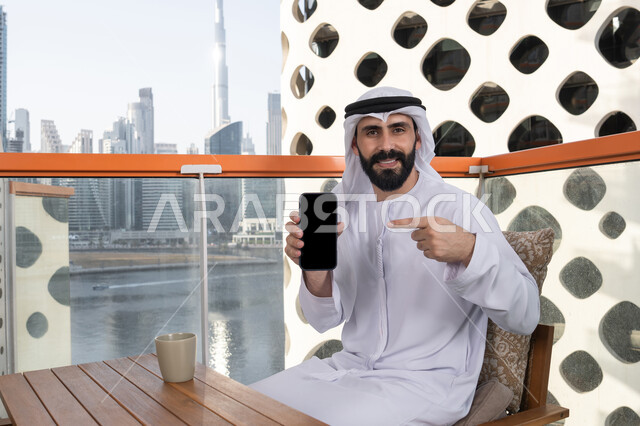 Looking at the camera with gestures of joy and happiness, displaying a blank black screen on the mobile phone, using modern technological means, a smiling Emirati Gulf Arab man wearing a kandura and ghutrah sitting in a café holding the mobile phone and pointing at it, spending enjoyable time outdoors.