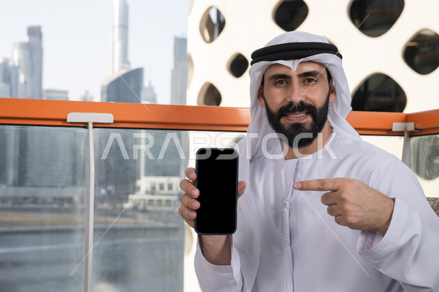 Using modern technological means, displaying a blank black screen on the mobile phone, looking at the camera with gestures of joy and happiness, a smiling Emirati Gulf Arab man wearing a kandura and ghutrah sitting in a café holding the mobile phone and pointing to it, spending enjoyable time outdoors.