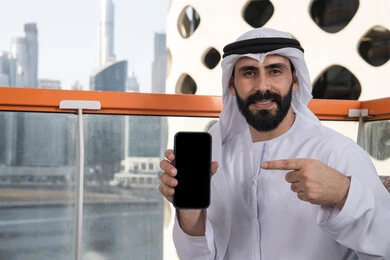 Using modern technological means, displaying a blank black screen on the mobile phone, looking at the camera with gestures of joy and happiness, a smiling Emirati Gulf Arab man wearing a kandura and ghutrah sitting in a café holding the mobile phone and pointing to it, spending enjoyable time outdoors.