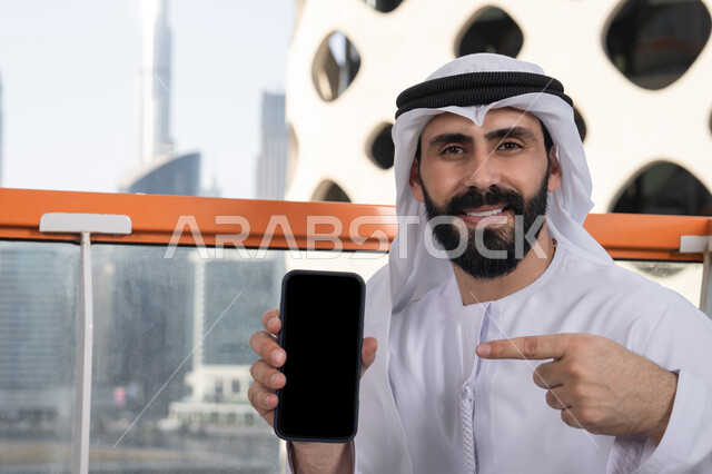 A black empty screen displayed on the mobile phone, using modern technological means, looking at the camera with gestures of joy and happiness, a close-up image of a smiling Emirati Gulf Arab man wearing a kandura and ghutrah sitting in a café holding the mobile phone and pointing to it, spending enjoyable time outdoors.