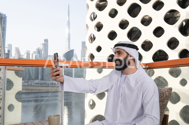 اجراء مكالمه فيديو, الاحتفاظ باللحظات السعيدة، التقاط صورة سيلفي تذكارية عبر جهاز الجوال بإيماءات السعادة والسرور، صورة من الجانب لرجل عربي خليجي اماراتي يرتدي الكندورة و الغتره يجلس فى المقهي يمسك بيده الهاتف المحمول، استخدام جهاز تقني حديث ومتطور