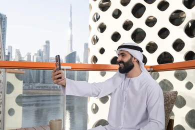 اجراء مكالمه فيديو, الاحتفاظ باللحظات السعيدة، التقاط صورة سيلفي تذكارية عبر جهاز الجوال بإيماءات السعادة والسرور، صورة من الجانب لرجل عربي خليجي اماراتي يرتدي الكندورة و الغتره يجلس فى المقهي يمسك بيده الهاتف المحمول، استخدام جهاز تقني حديث ومتطور