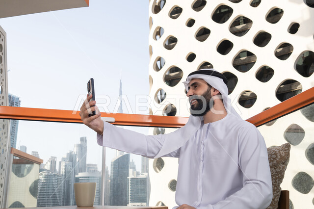 التقاط صور السيلفي التذكارية عبر الجوال, ايماءات السعادة و السرور, صورة من الجانب لرجل عربي خليجي اماراتي يرتدي الكندورة و الغتره يجلس فى المقهي يمسك بيده الهاتف المحمول، استخدام جهاز تقني حديث ومتطور