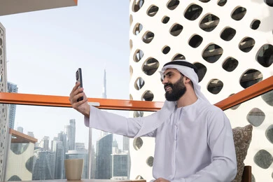 Taking commemorative selfie photos via mobile, gestures of happiness and joy, a side image of an Emirati Gulf Arab man wearing a kandura and ghutrah sitting in a café holding a mobile phone in his hand, using a modern and advanced technological device.