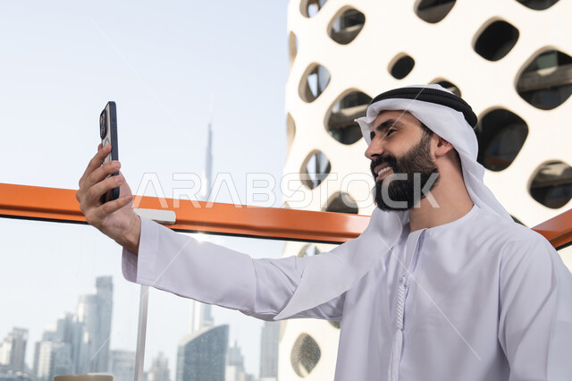 تصوير اللحظات السعيدة، التقاط صورة سيلفي تذكارية عبر جهاز الجوال بإيماءات السعادة والسرور، تصوير مقرب من الجانب لرجل عربي خليجي اماراتي يرتدي الكندورة و الغتره يجلس فى المقهي يمسك بيده الهاتف المحمول، استخدام جهاز تقني حديث ومتطور