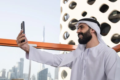 Capturing happy moments, taking a commemorative selfie with a mobile device with gestures of happiness and joy, a close-up side shot of an Emirati Gulf Arab man wearing a kandura and ghutrah sitting in a café holding a mobile phone in his hand, using a modern and advanced technological device.