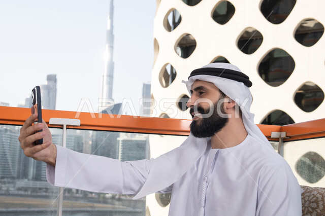 الاحتفاظ بالذكريات السعيدة، التقاط صورة سيلفي عبر جهاز الجوال بإيماءات السعادة والسرور،صورة  مقربة من الجانب رجل عربي خليجي اماراتي يرتدي الكندورة و الغتره يجلس فى المقهي يمسك بيده الهاتف المحمول، استخدام جهاز تقني حديث ومتطور