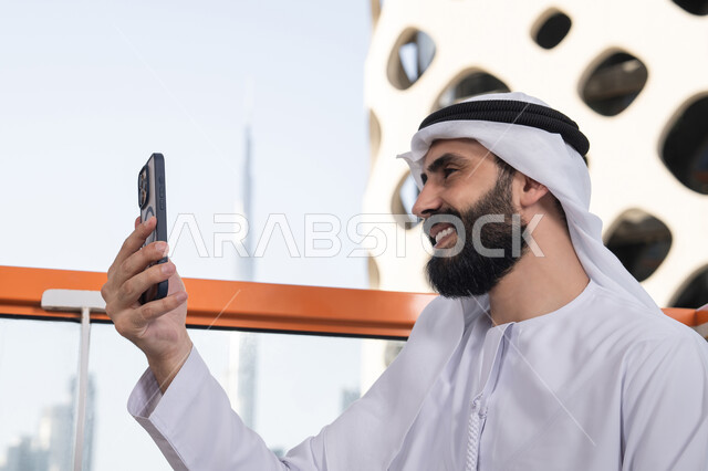 استخدام الجوال فى توثيق اللحظات التذكاريه,الاحتفاظ باللحظات السعيدة، إيماءات السعادة والسرور، صورة مقربة من الجانب لرجل عربي خليجي اماراتي يرتدي الكندورة و الغتره يجلس فى المقهي يمسك بيده الهاتف المحمول، استخدام جهاز تقني حديث ومتطور