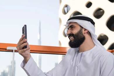 استخدام الجوال فى توثيق اللحظات التذكاريه,الاحتفاظ باللحظات السعيدة، إيماءات السعادة والسرور، صورة مقربة من الجانب لرجل عربي خليجي اماراتي يرتدي الكندورة و الغتره يجلس فى المقهي يمسك بيده الهاتف المحمول، استخدام جهاز تقني حديث ومتطور